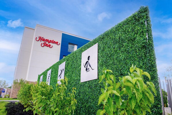 A Hampton Inn building with a blue facade and a green hedge wall.