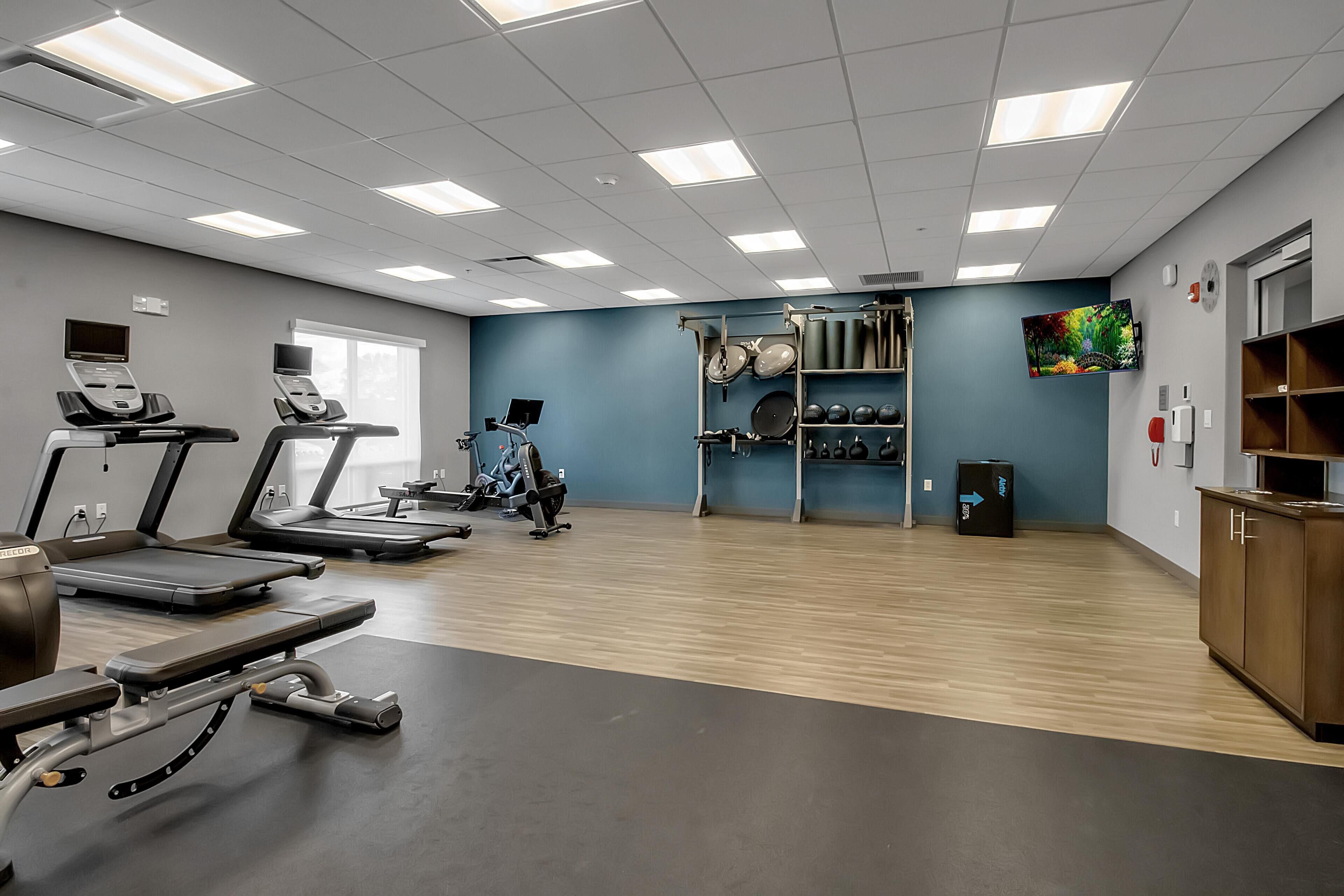 A gym with treadmills, a stationary bike, and a rowing machine. There is a wall-mounted TV and a shelf with exercise equipment.
