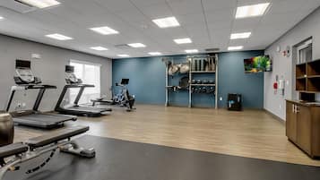 A gym with treadmills, a stationary bike, and a rowing machine. There is a wall-mounted TV and a shelf with exercise equipment.