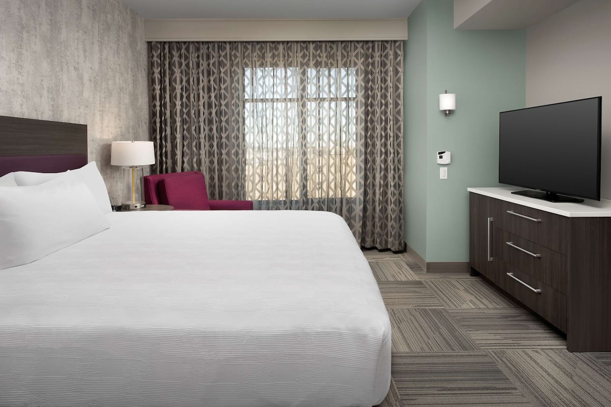 Una camera d'hotel con un letto grande, una TV su un comò, una poltrona rossa e una finestra con tende a motivi.