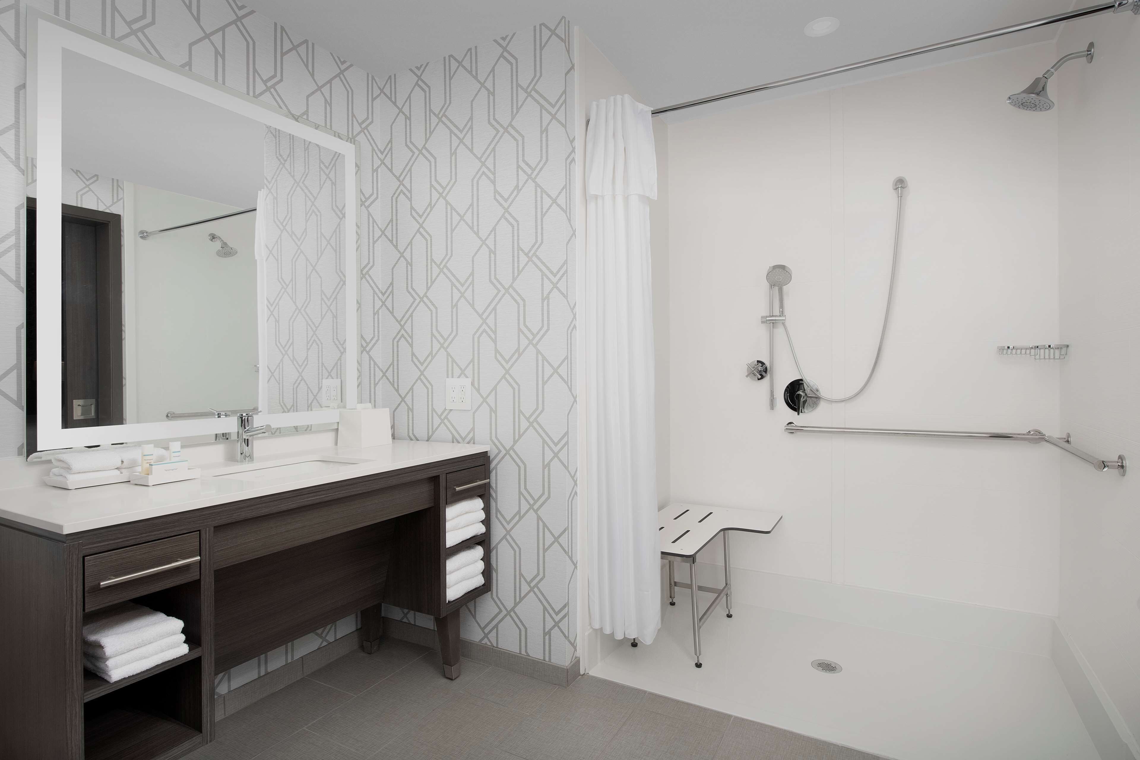 A modern bathroom with a large mirror, dark wooden vanity, white sink, and a walk-in shower with a handheld showerhead.