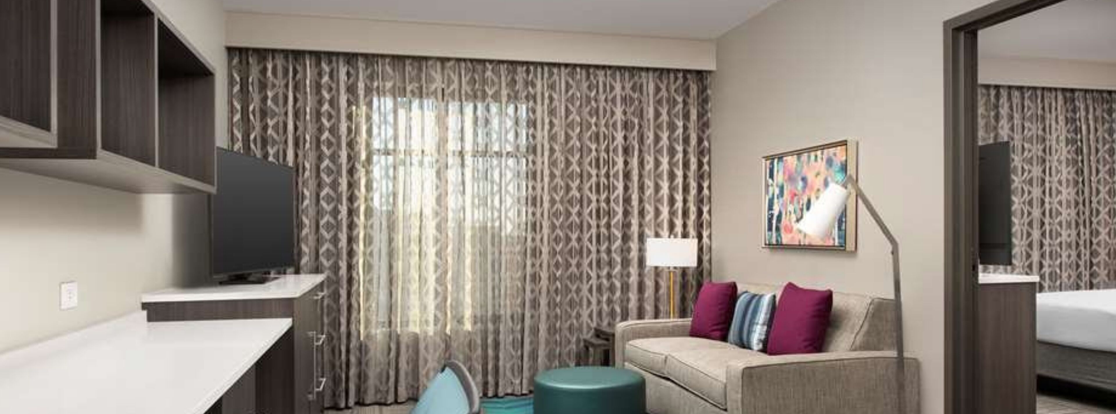 A modern hotel room with a flat-screen TV, a sofa with cushions, a desk, and a large window with patterned curtains.