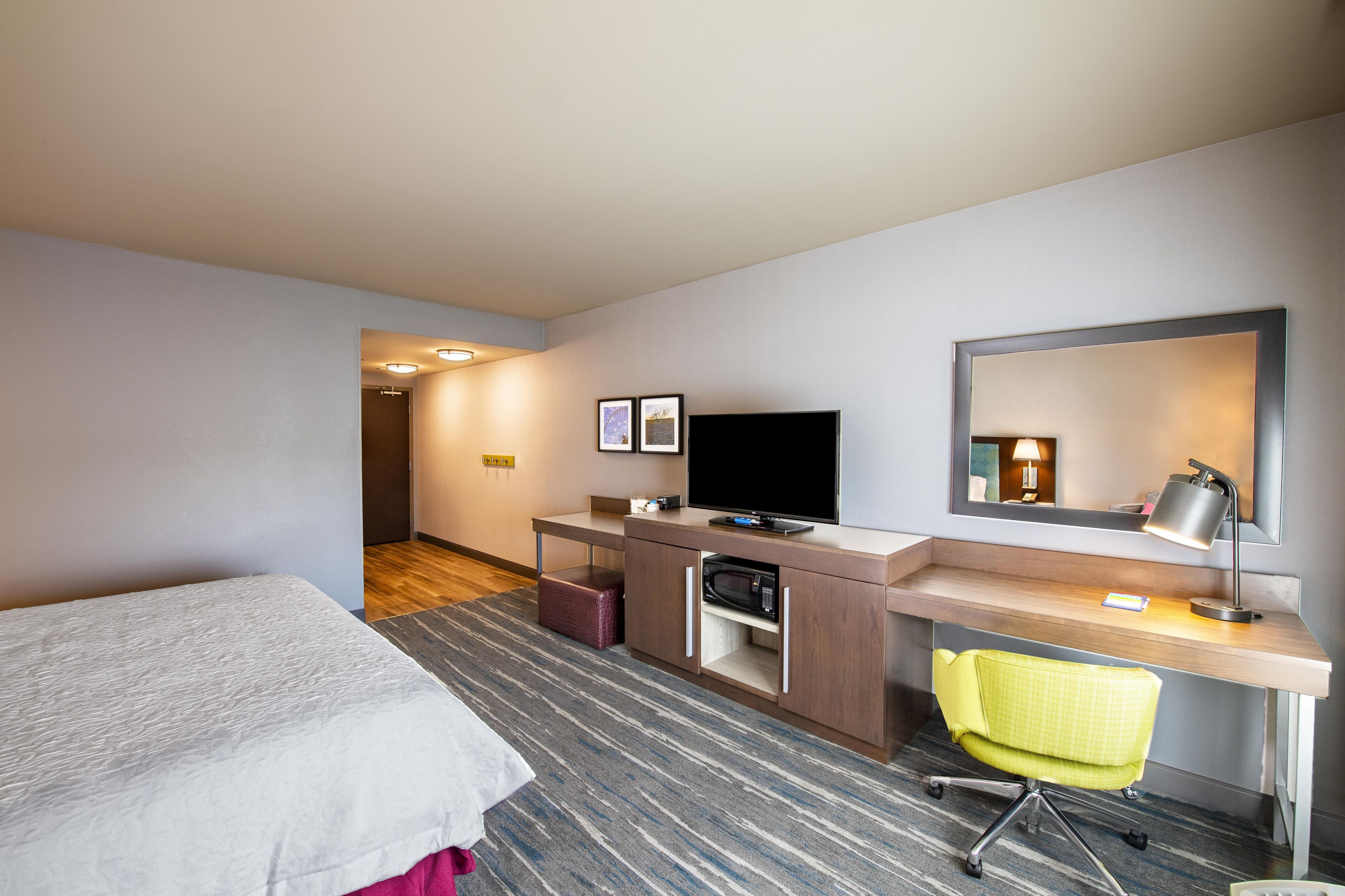 Una habitación de hotel con cama, escritorio, silla, televisor y espejo.