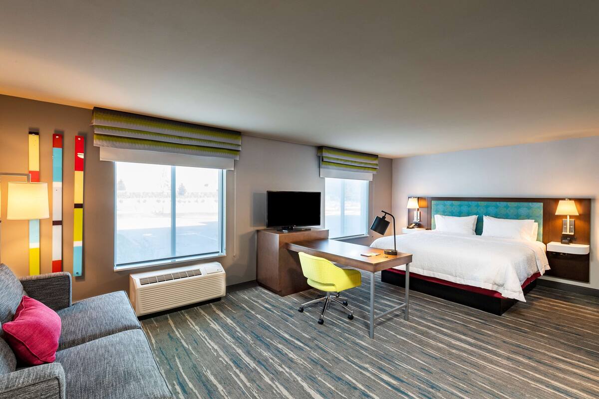 A hotel room with a bed, desk, chair, TV, and large windows.