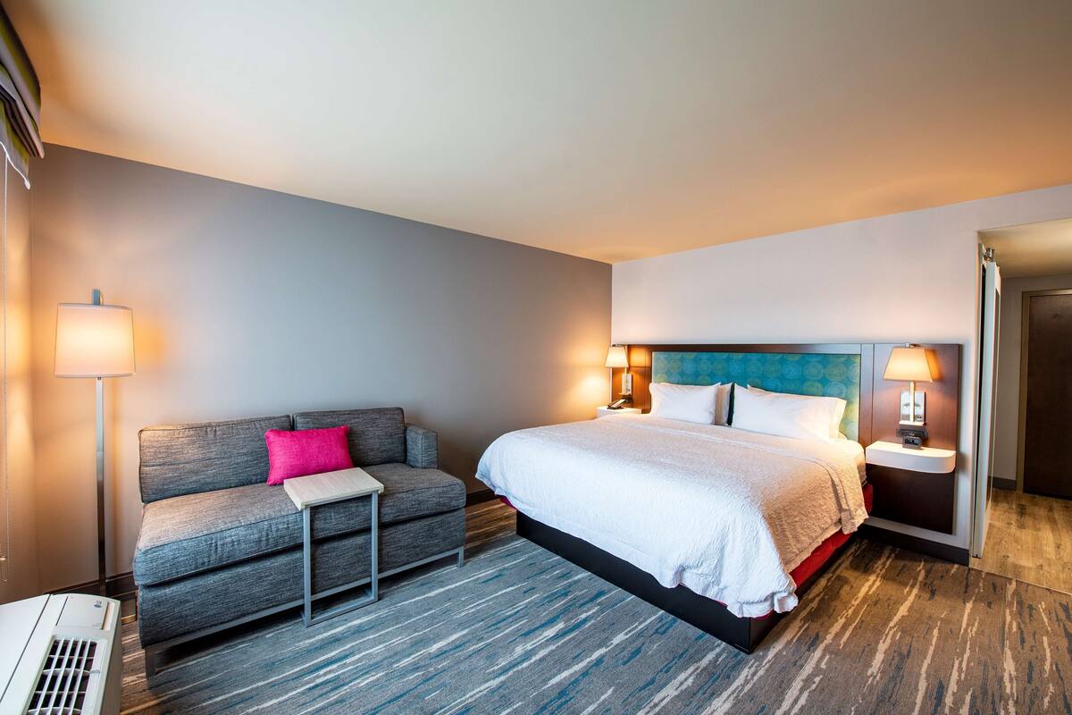 A hotel room with a bed, a sofa, a small table, and a lamp.