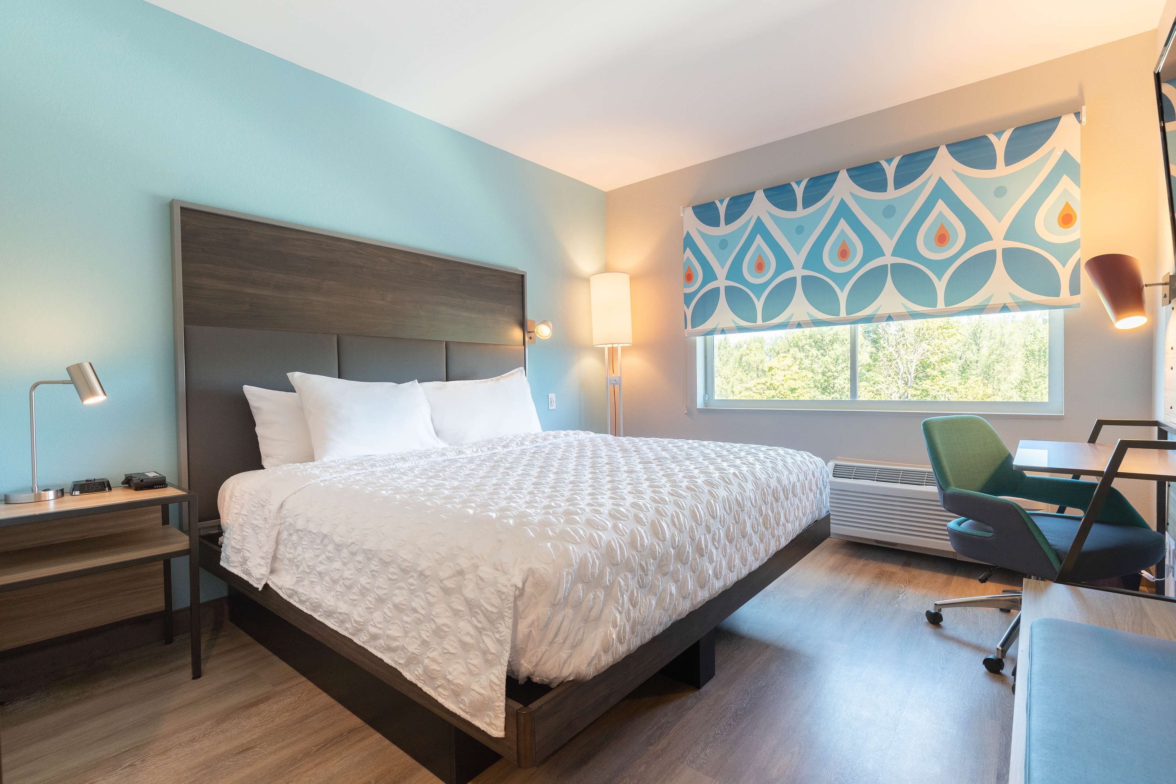 A modern hotel room with a large bed, a desk with a lamp, a chair, and a window with patterned curtains.