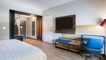 Quarto de hotel com cama, escrivaninha com cadeira, TV fixada na parede e porta para o banheiro.