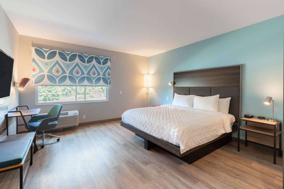 A modern hotel room with a large bed, a desk with a chair, a TV, and a window with patterned curtains.