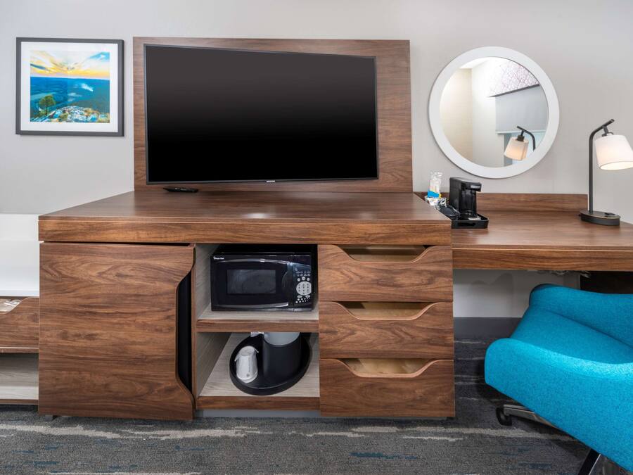 Habitación de hotel moderna con un escritorio de madera, un televisor de pantalla plana, un microondas, una cafetera, un espejo redondo y una silla azul.