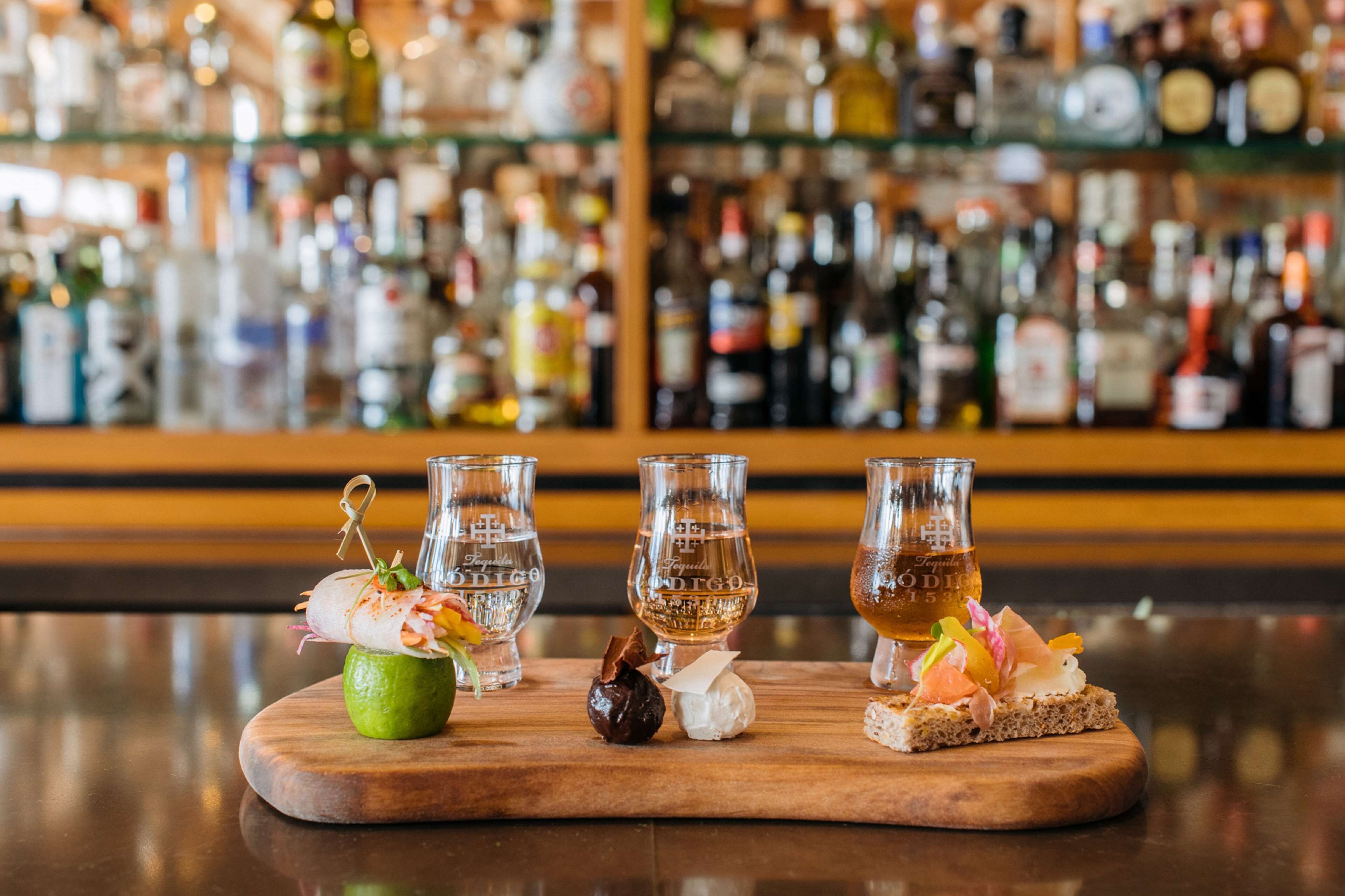 A bar counter with three glasses of liquor, a lime, and various garnishes.