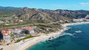 Un complejo turístico costero con varias edificaciones, una playa y terreno montañoso al fondo.