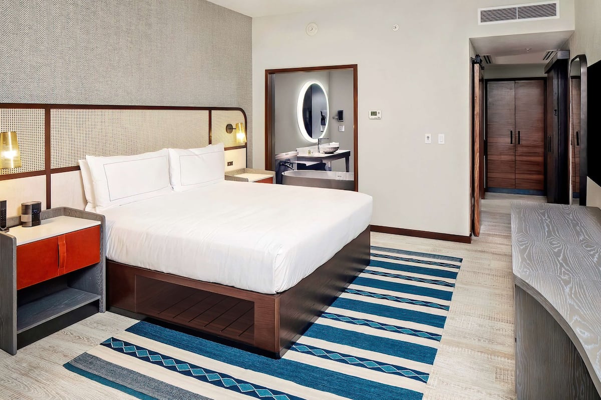 Habitación de hotel moderna con una cama grande, mesitas de noche, un espejo y un baño visible a través de una puerta abierta.