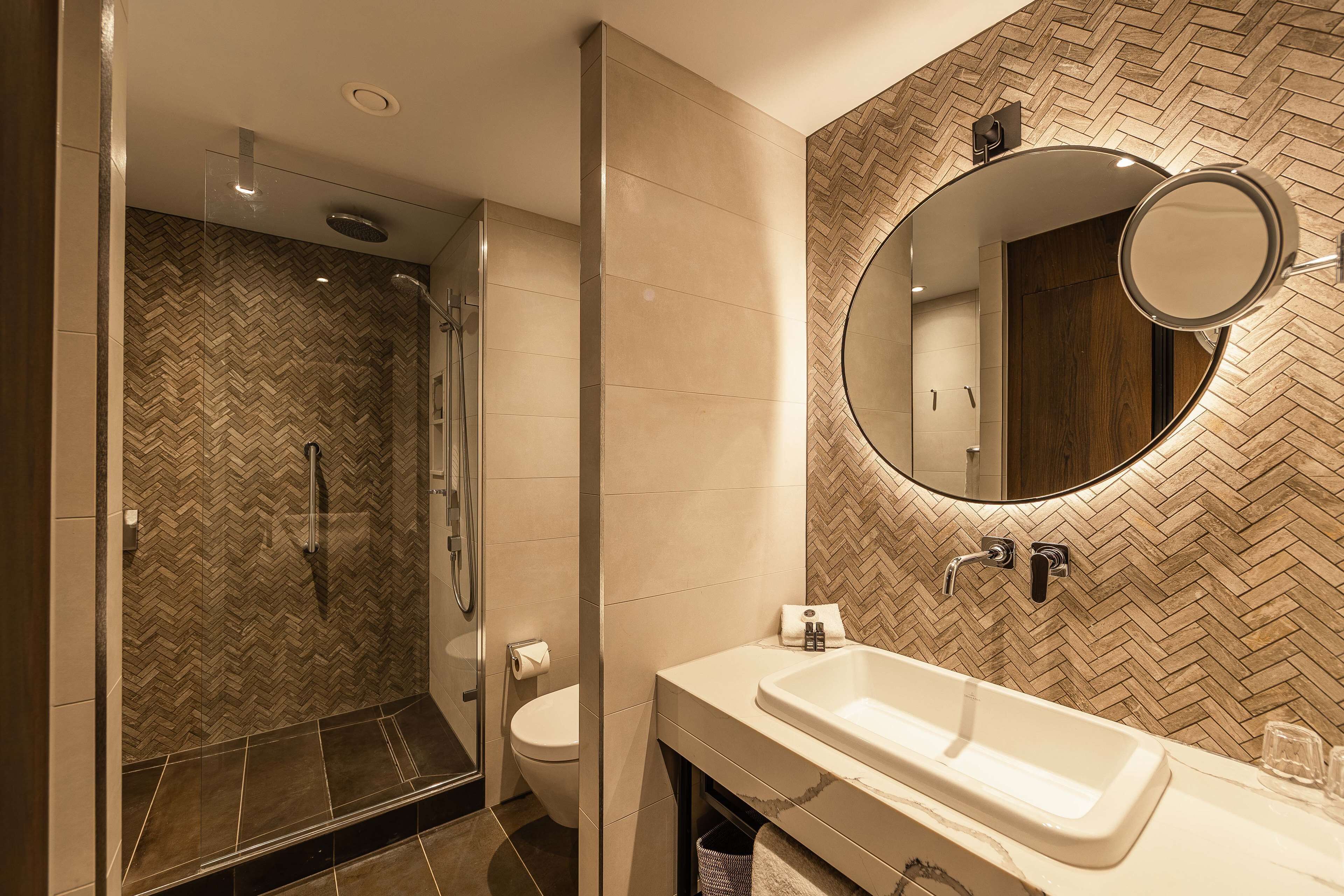 Un baño moderno con pared de diseño en patrón espiga, ducha con cerramiento de vidrio, lavamanos blanco y espejo redondo.