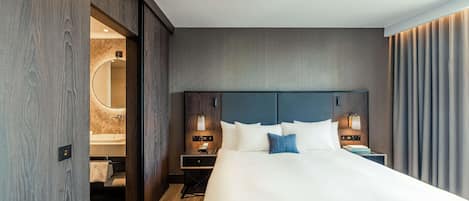 A modern hotel room with a large bed, a wooden wardrobe, a bedside table with a lamp, and a bathroom visible through an open door.