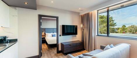 Habitación de hotel moderna con suelo de madera, sofá, televisión, mesita de noche y vistas al exterior.
