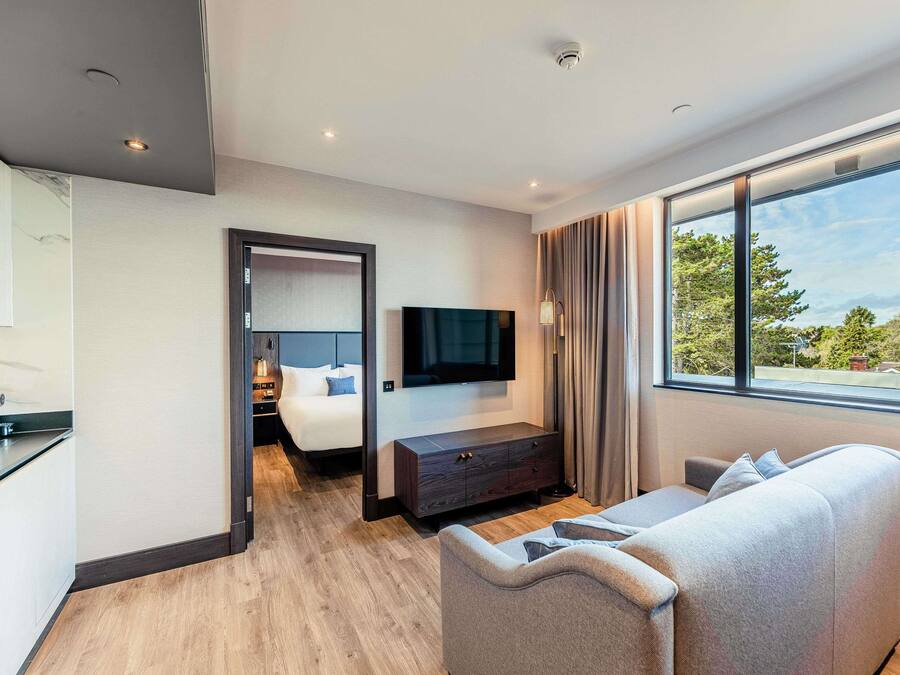 A modern hotel room with a wooden floor, a sofa, a TV, a bedside table, and a view of the outdoors.