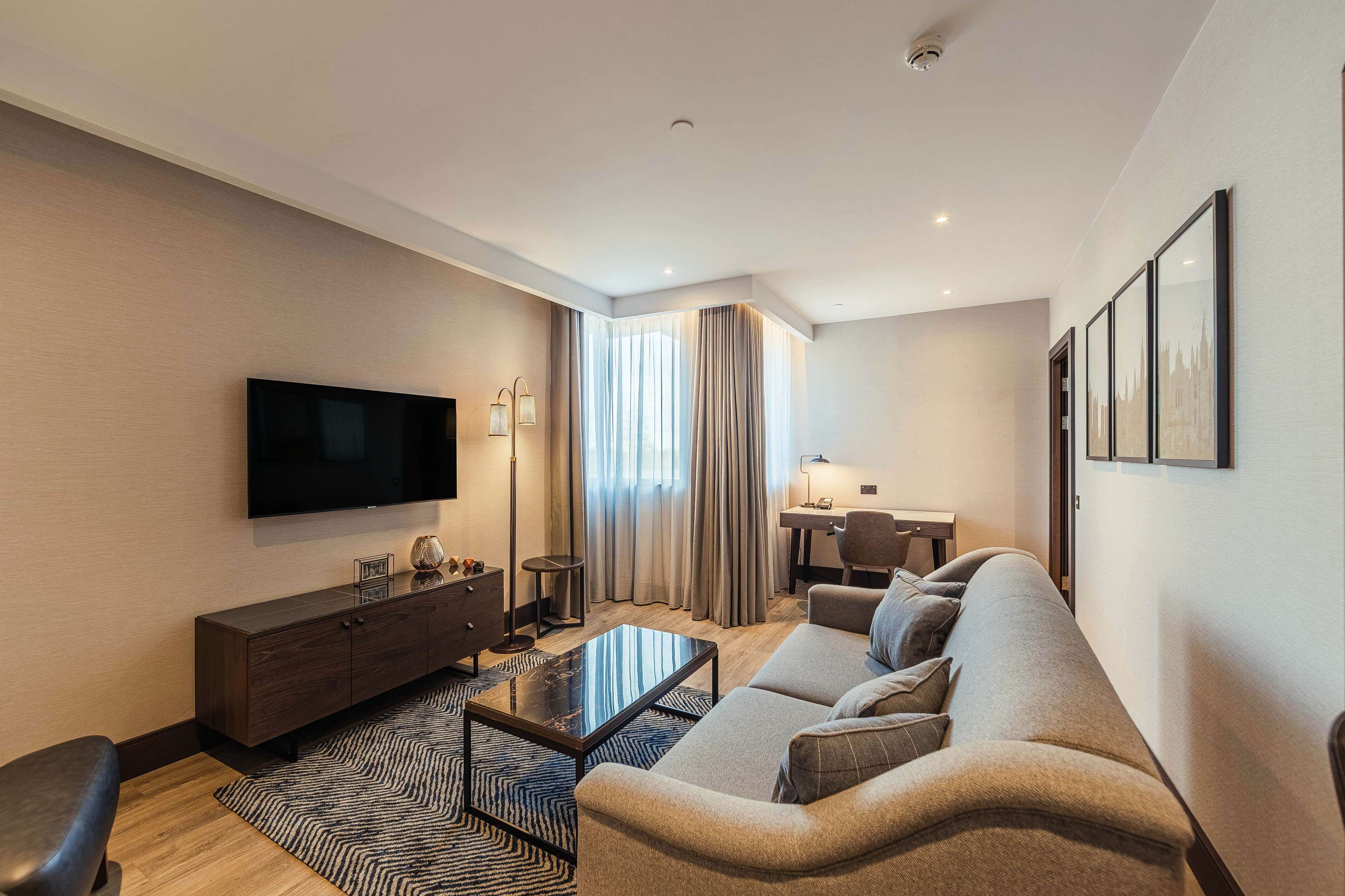 A modern living room with a flat-screen TV, a sofa, a coffee table, and a desk with a lamp.