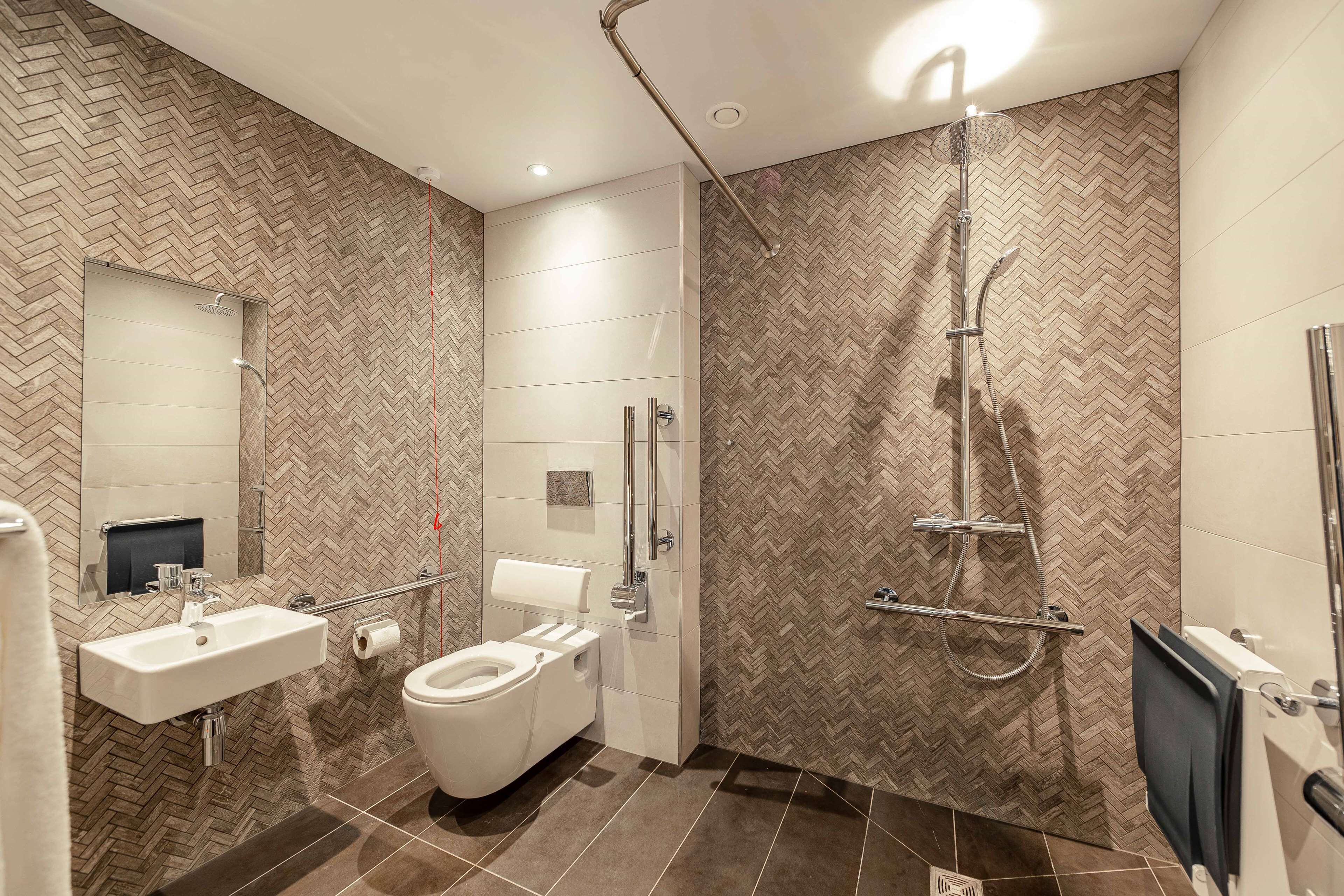 A modern bathroom with a herringbone-patterned wall, a white sink, a toilet, and a walk-in shower with a handheld showerhead.