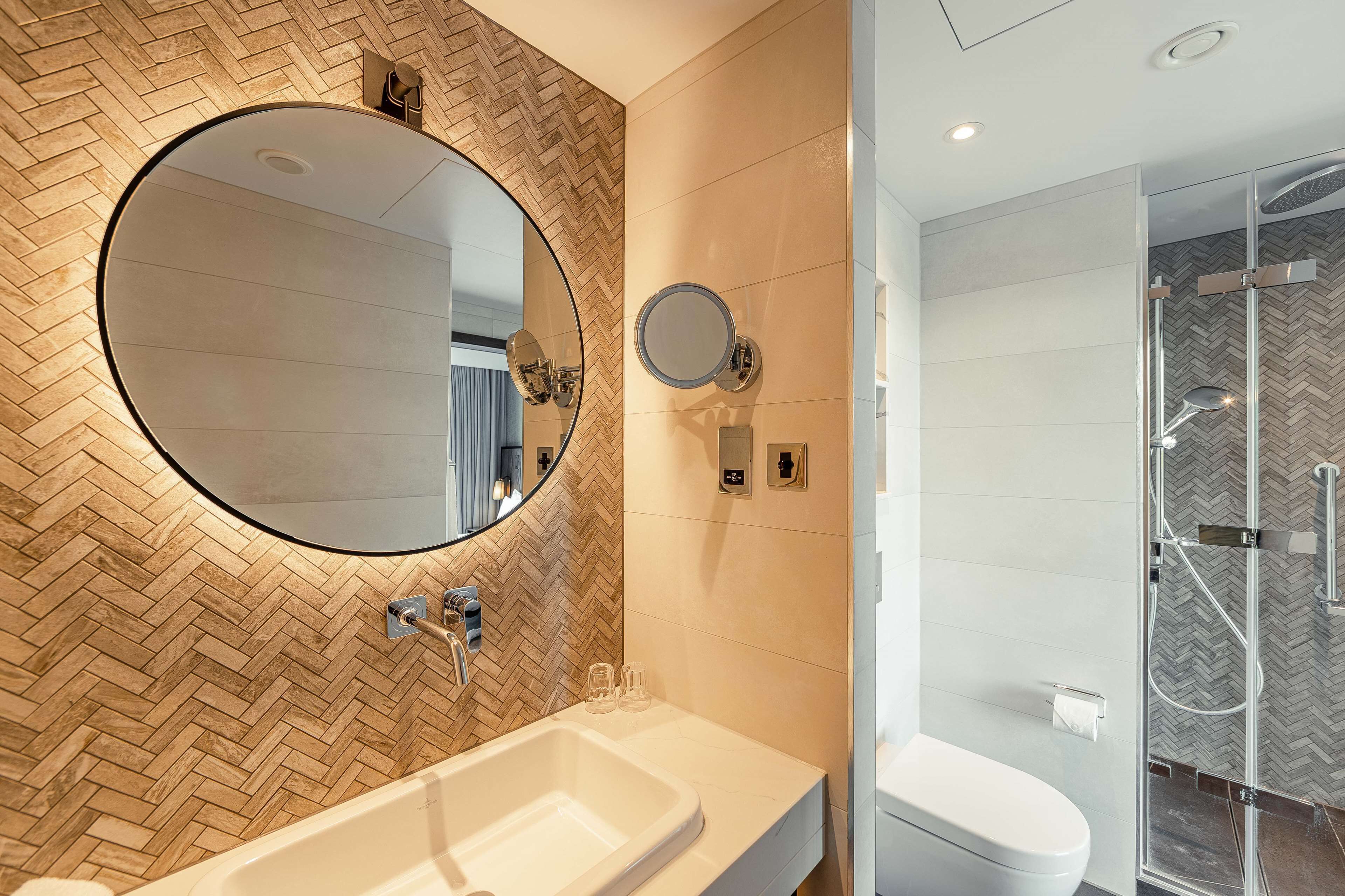 Une salle de bain moderne avec un mur recouvert de carreaux disposés en motif chevron, un grand miroir rond, un lavabo blanc et une douche à parois de verre.