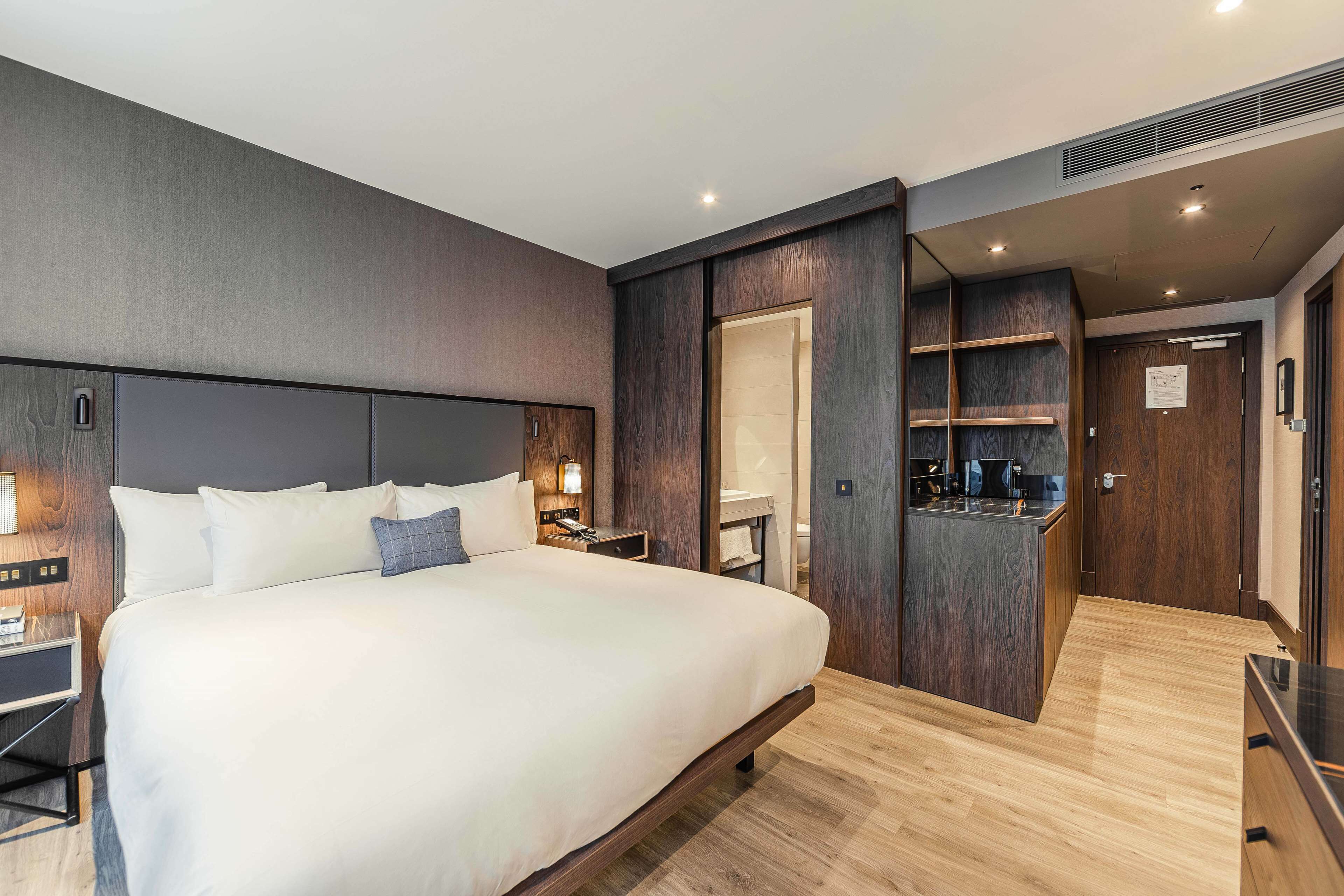 Une chambre d’hôtel moderne dotée d’un grand lit, de tables de chevet, d’un bureau intégré et d’une petite kitchenette.