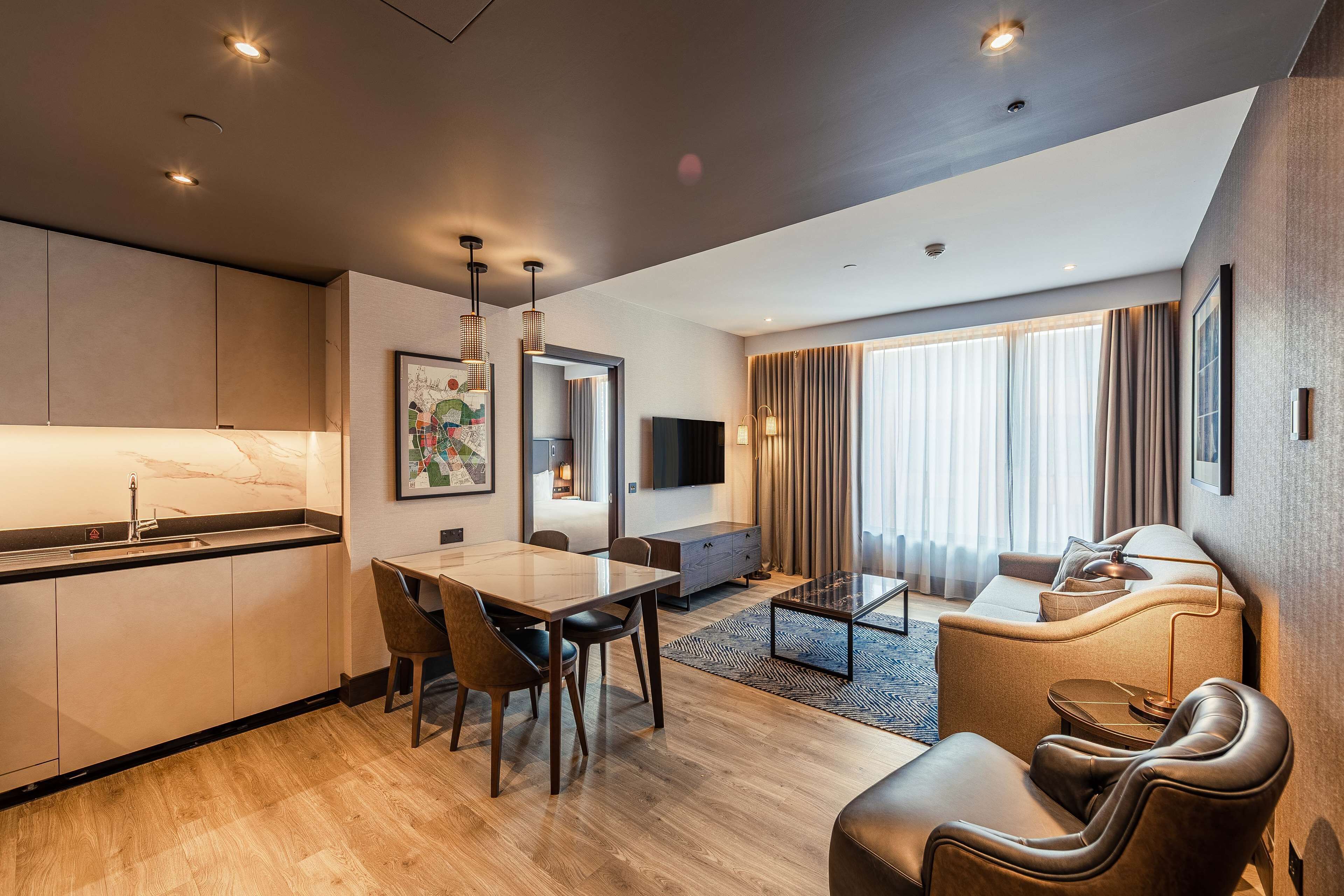 A modern hotel room with a dining area, a sofa, a TV, and a large window with curtains.