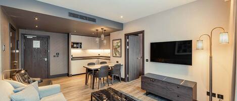 A modern living room with a sofa, a coffee table, and a TV mounted on the wall. There is a dining area with a table and chairs, and a kitchen in the background.