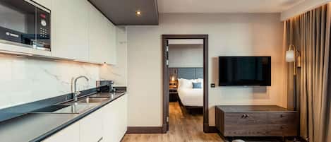 A modern hotel room with a kitchen area, a bed, a TV, and a seating area.