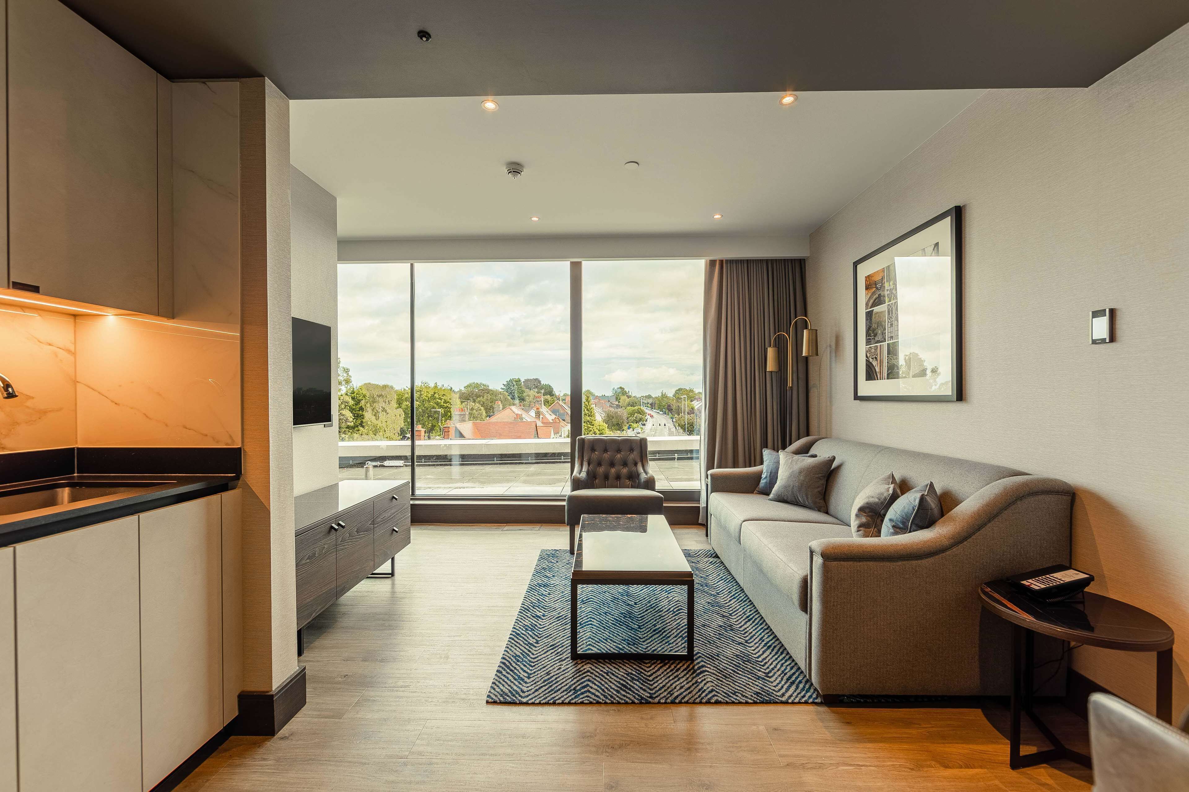A modern living room with a sofa, a coffee table, and a TV. There is a kitchen area with a marble countertop and cabinets. The room has large windows with a view of the city.