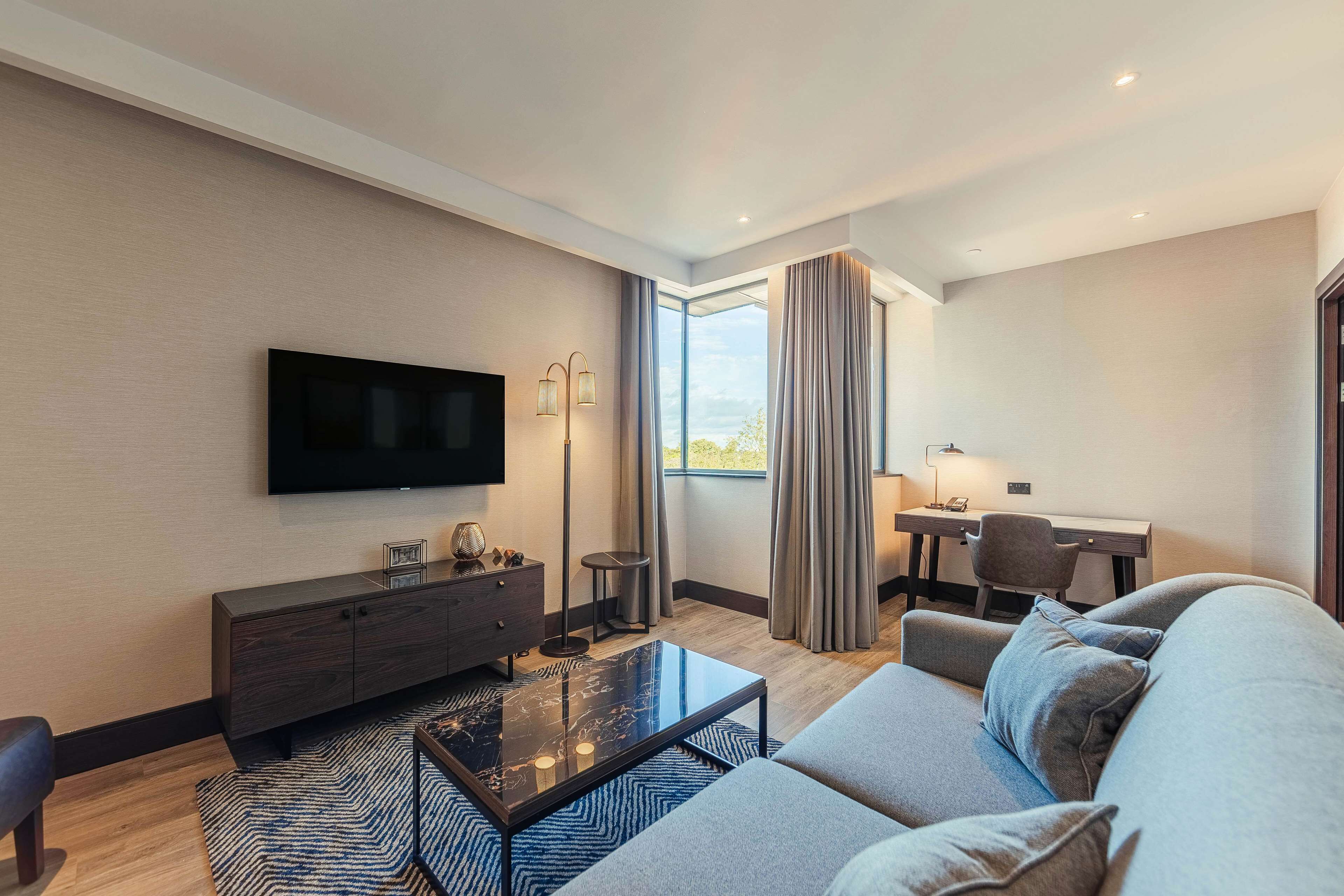 Een moderne hotelkamer met een flatscreen televisie, een bank, een salontafel, een bureau met stoel, en een groot raam met gordijnen.