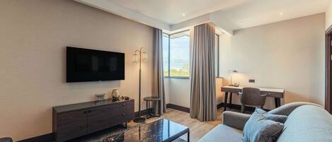 Een moderne hotelkamer met een flatscreen televisie, een bank, een salontafel, een bureau met stoel, en een groot raam met gordijnen.