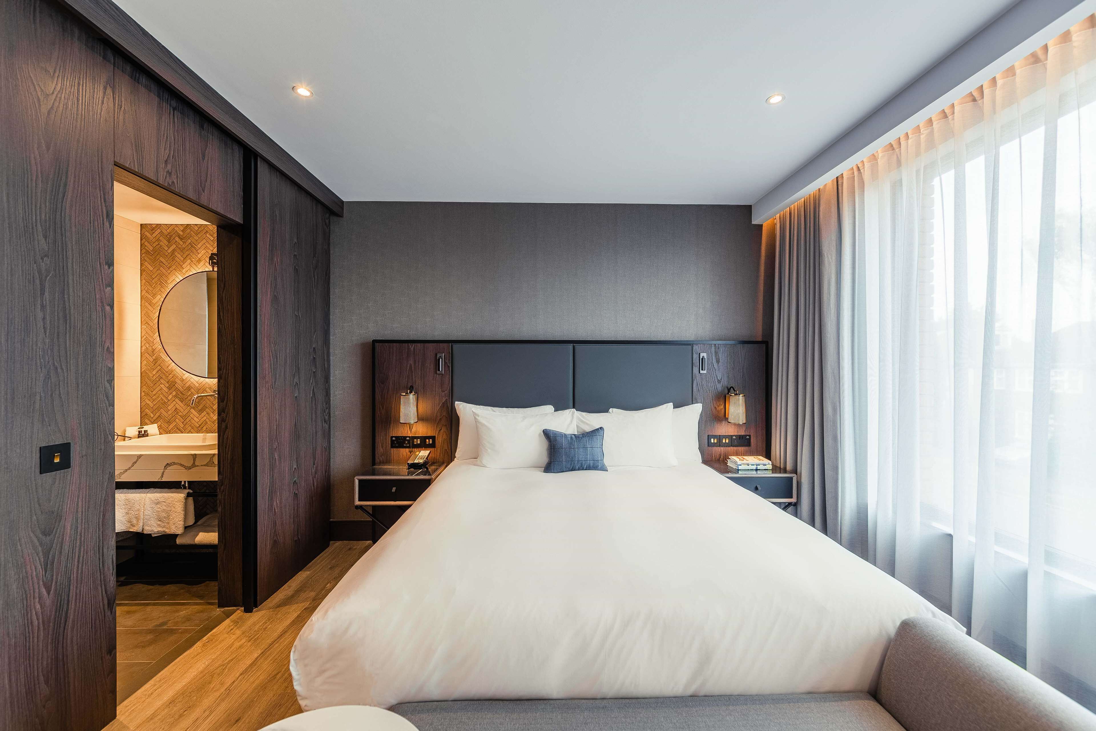 A modern hotel room with a large bed, wooden paneling, a vanity with a mirror, and a view of the outdoors through sheer curtains.