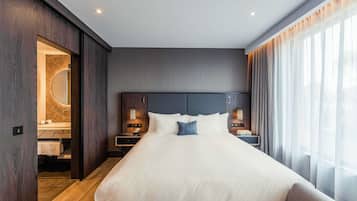 A modern hotel room with a large bed, wooden paneling, a vanity with a mirror, and a view of the outdoors through sheer curtains.