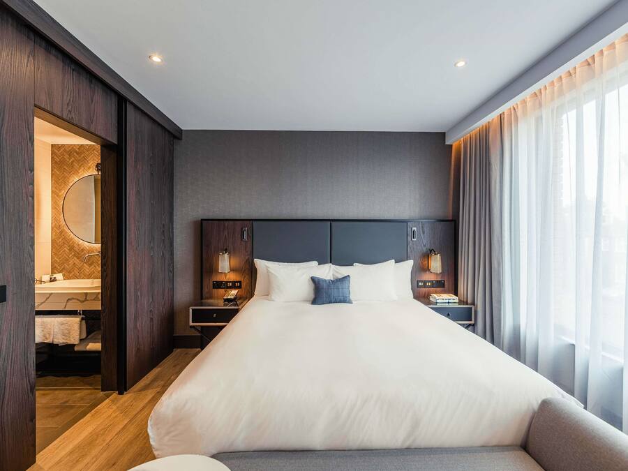 A modern hotel room with a large bed, wooden paneling, a vanity with a mirror, and a view of the outdoors through sheer curtains.