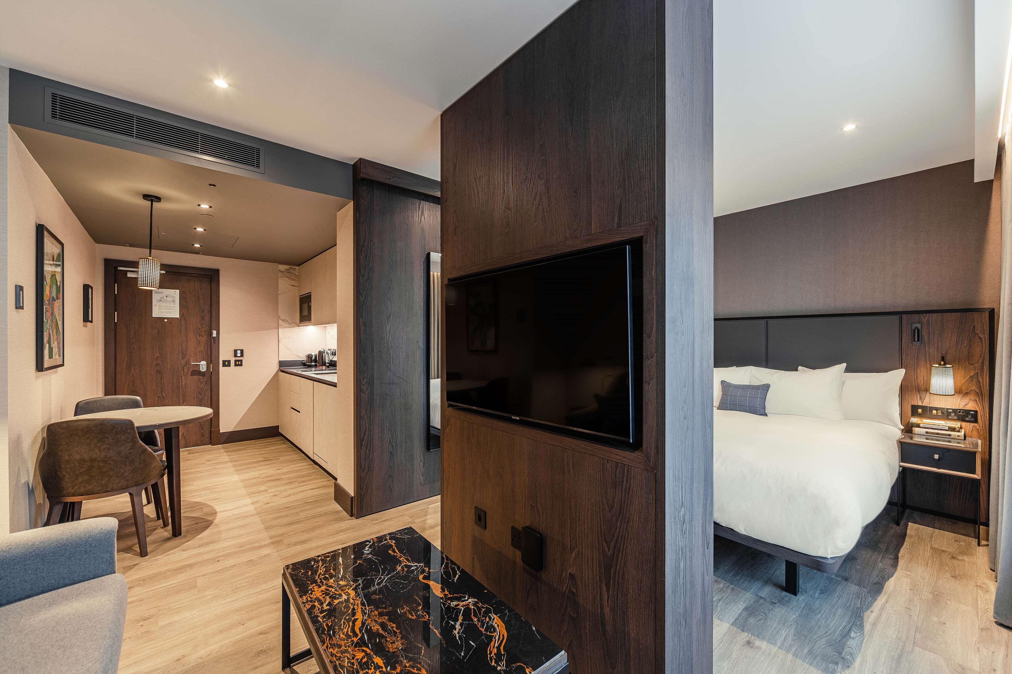 A modern hotel room with a large bed, a TV mounted on the wall, a dining area with a table and chairs, and a kitchen area.