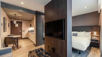 A modern hotel room with a large bed, a TV mounted on the wall, a dining area with a table and chairs, and a kitchen area.