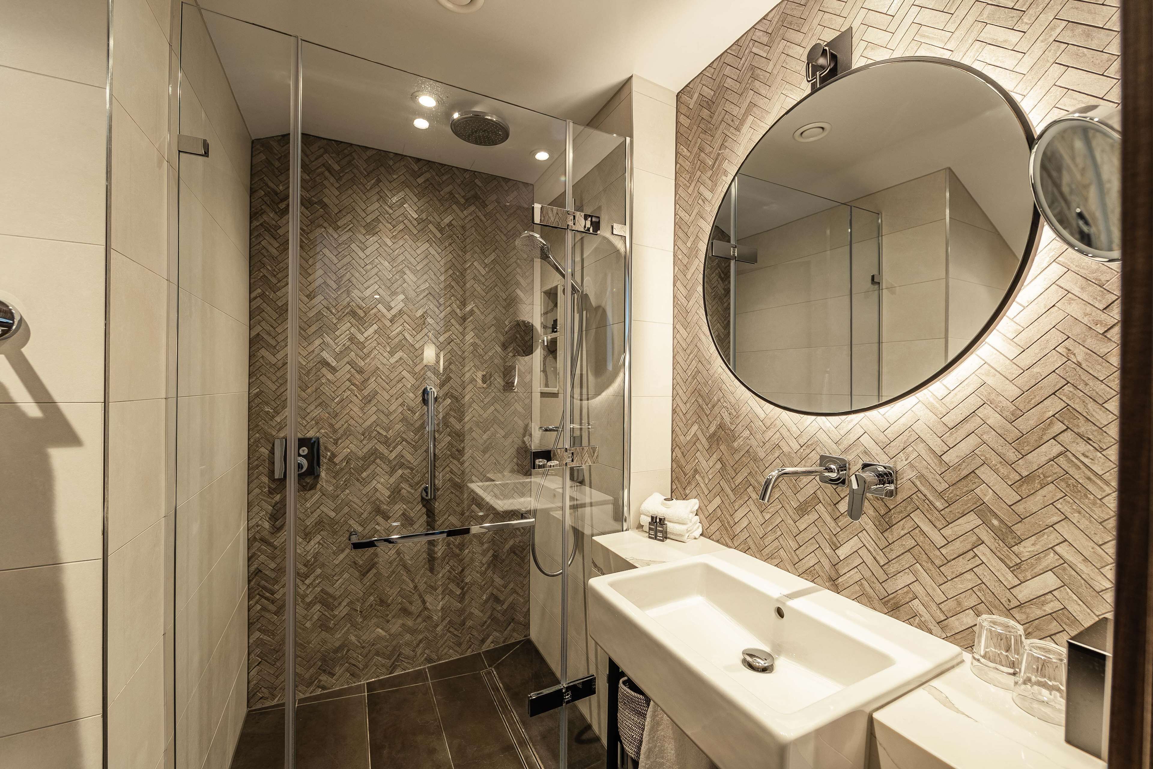 A modern bathroom with a glass-enclosed shower, a large mirror, and a herringbone-patterned tile backsplash.