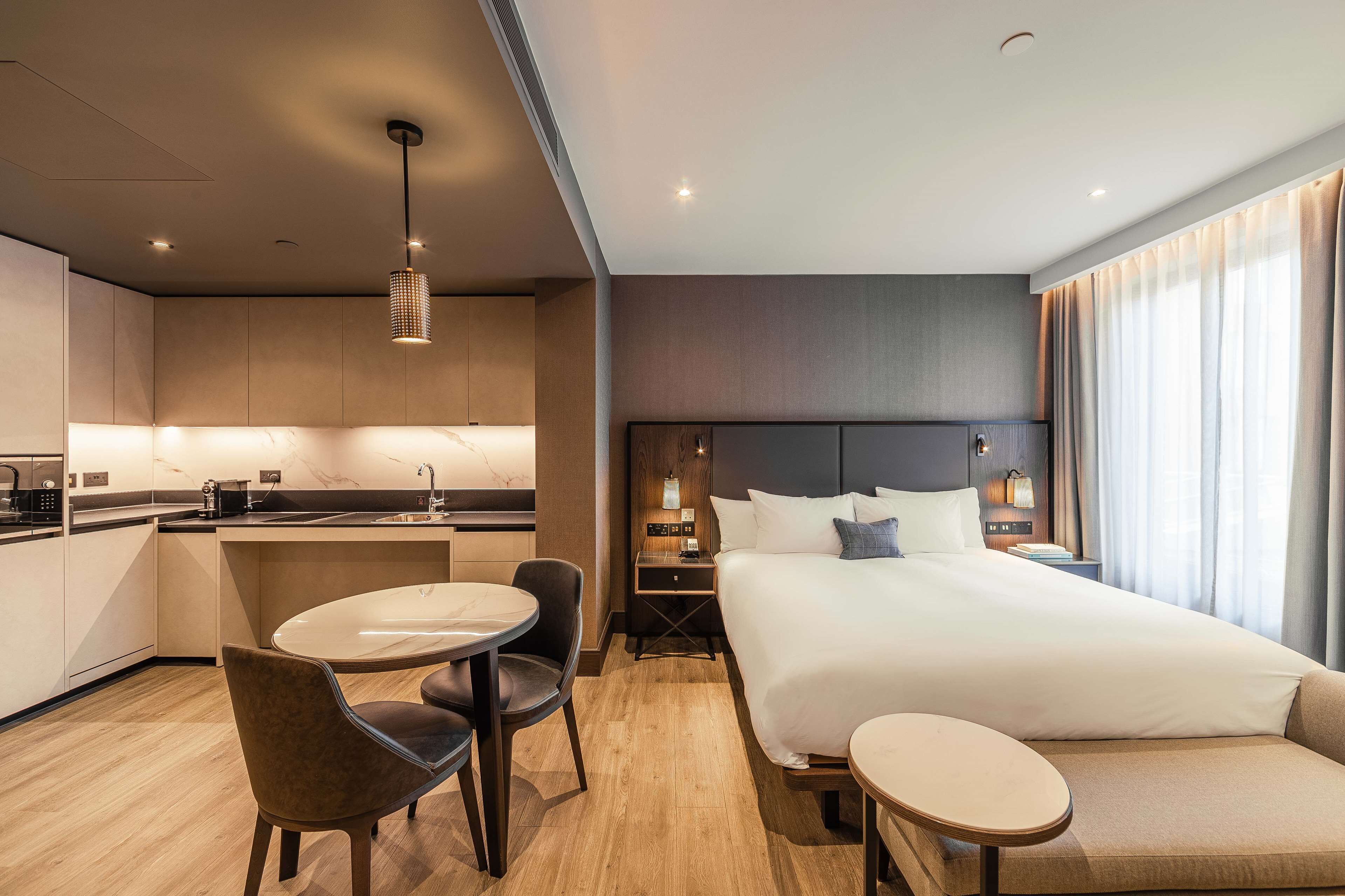 Una habitación de hotel moderna con una cama grande, una pequeña mesa de comedor, una zona de estar separada y una kitchenette.