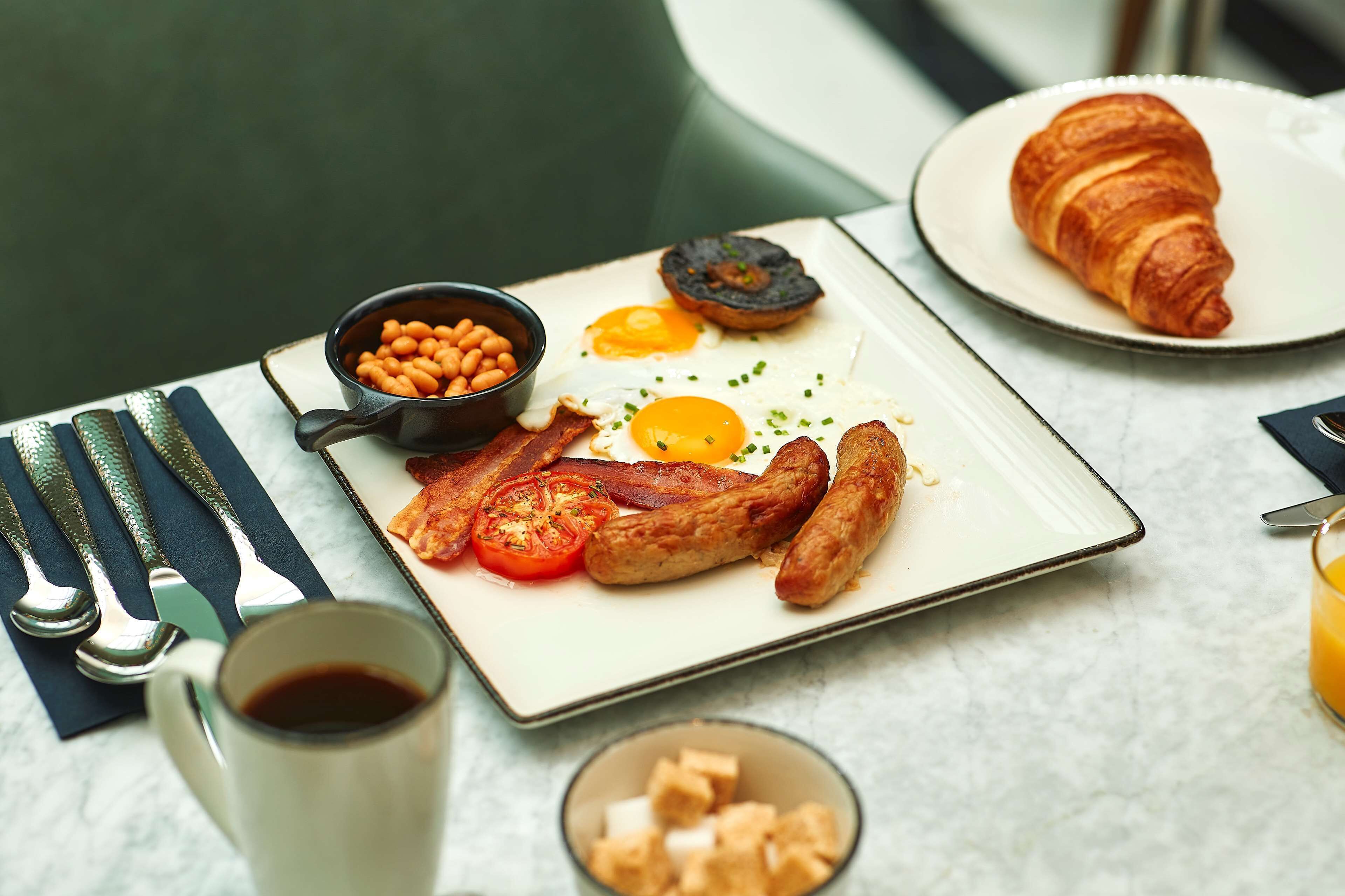 Un plato de desayuno con huevos, tocino, salchichas, tomates y frijoles, acompañado de un croissant y una taza de café.