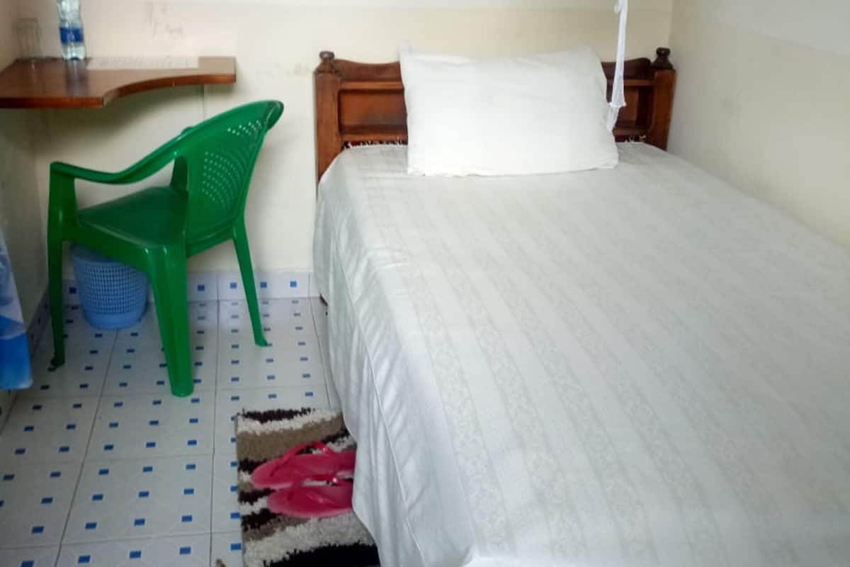 A single bed with a white bedspread, a wooden headboard, a green plastic chair, a small wooden table with a bottle, and a white hanging light fixture.