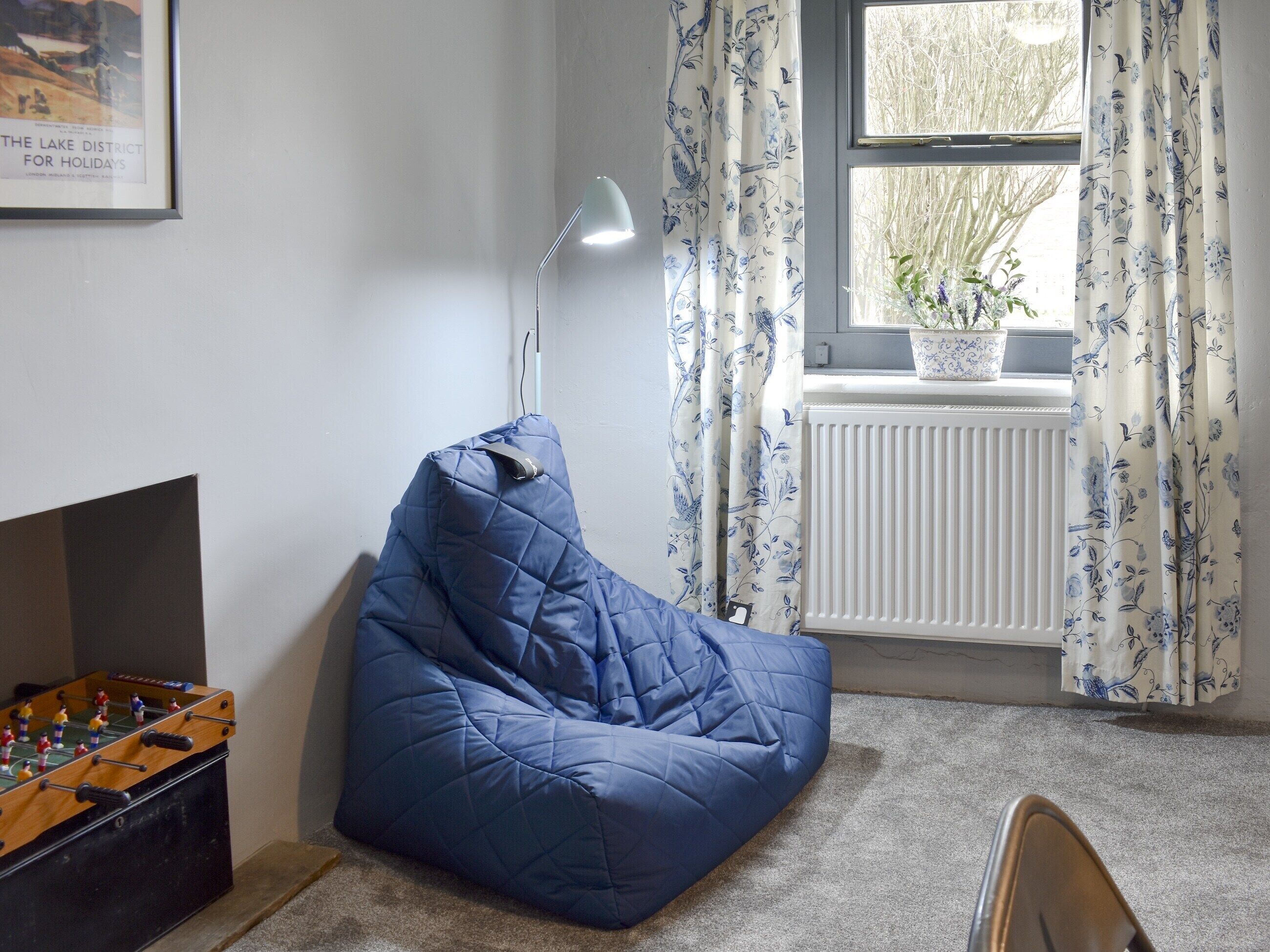 Une pièce avec un fauteuil en forme de sac de haricots bleu, un baby-foot, un radiateur et des rideaux à motifs floraux.