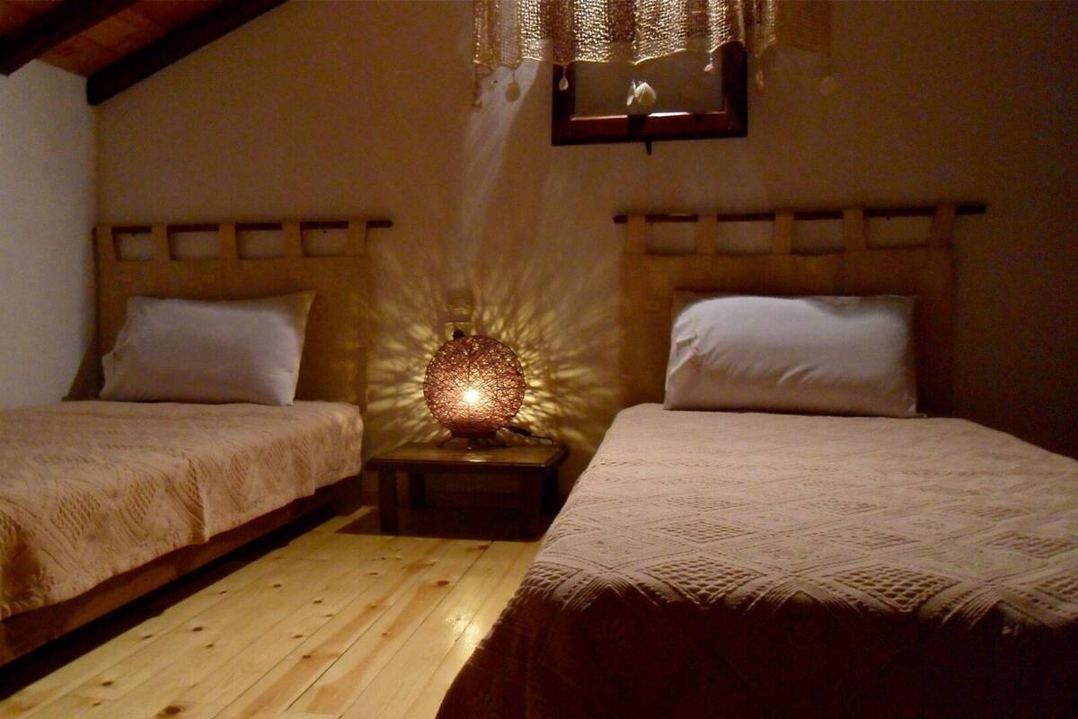 A cozy bedroom with two single beds, wooden headboards, a small bedside table with a lamp, and a window with a curtain.