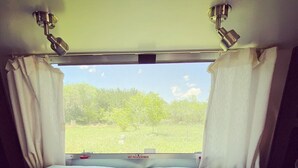 A bed with white bedding and two pillows, curtains on either side, and a window with a view of greenery outside. - Airstream at The Goat (San Marcos)
