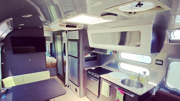 A compact kitchen with a sink, stove, and refrigerator. There is a dining area with a bench and a small table. - Airstream at The Goat (San Marcos)
