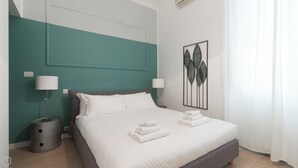 A neatly made bed with white linens and towels, a teal accent wall, a modern lamp, and a leaf-patterned artwork on the wall.