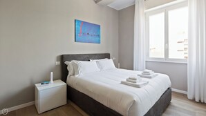 A neatly made bed with white linens and a gray headboard, a bedside table with a no-smoking sign, and a window with white curtains. - Italianway - Largo Allegri 4 (Milan)