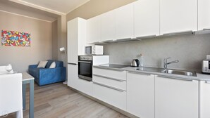 A modern kitchen with white cabinets, a microwave, a kettle, and a toaster. There is a blue sofa and a dining table with a wine glass. - Italianway - Largo Allegri 4 (Milan)