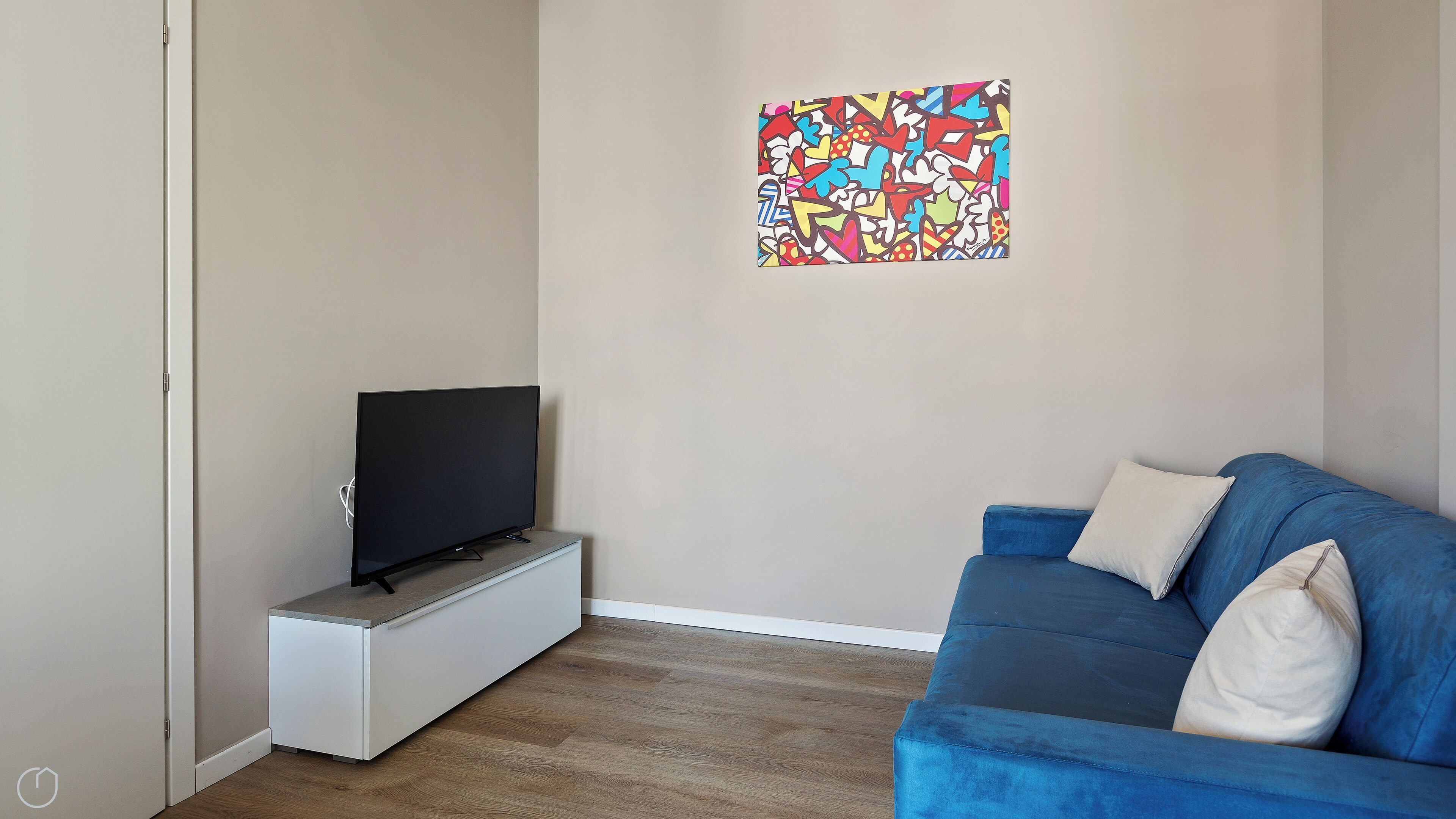 A living room with a flat-screen TV on a stand, a blue sofa with cushions, and a colorful abstract painting on the wall.