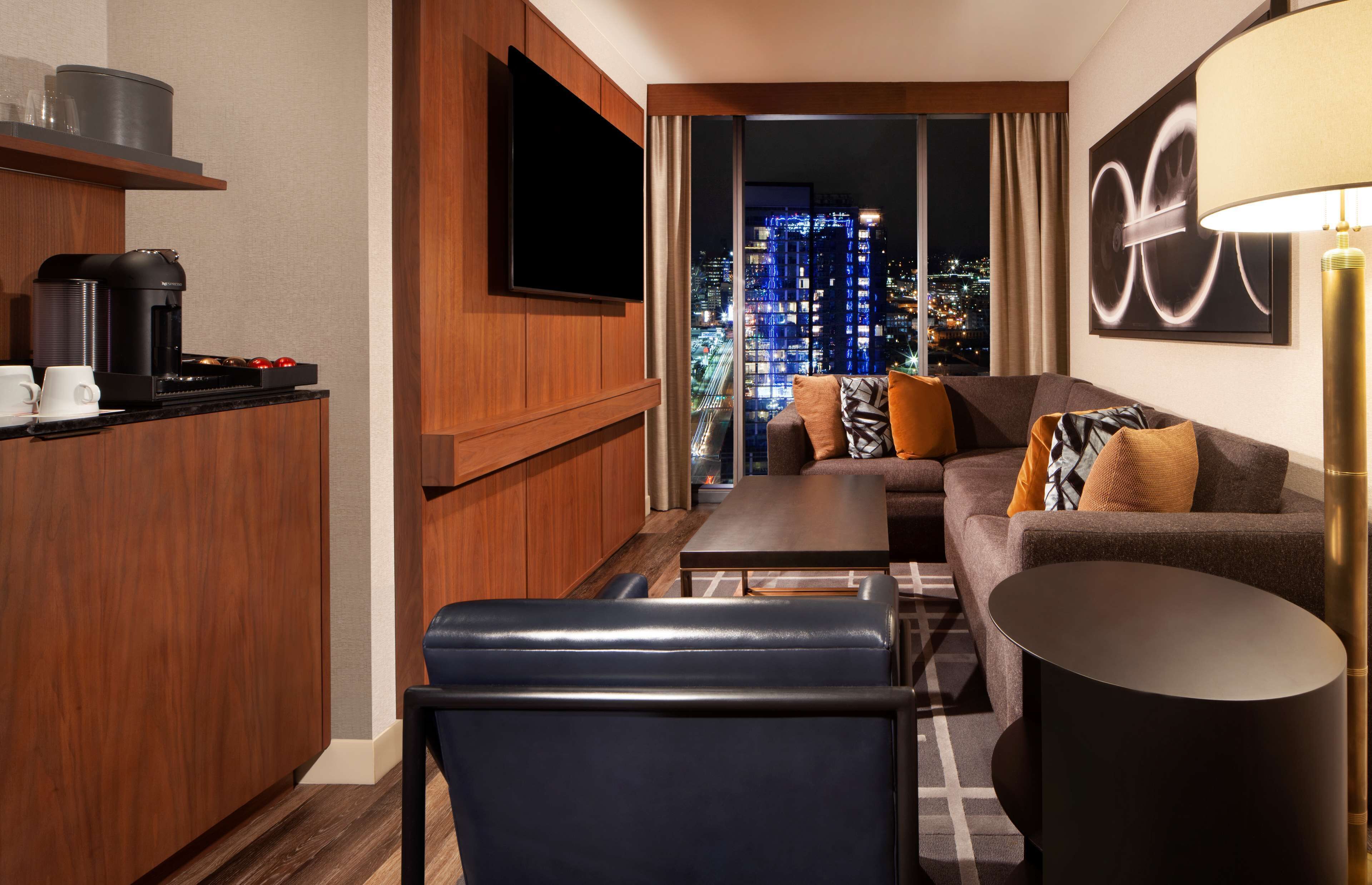 Habitación de hotel moderna con sala de estar, cocina equipada con cafetera, televisor de pantalla plana y vistas a la ciudad.