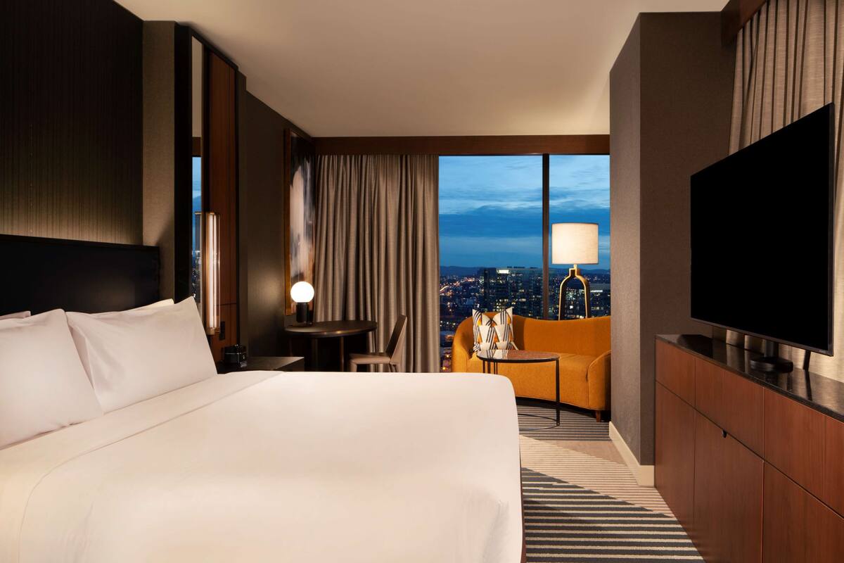 Een hotelkamer met een groot bed, een televisie, een bureau met lamp, een stoel en uitzicht op de stad 's nachts.