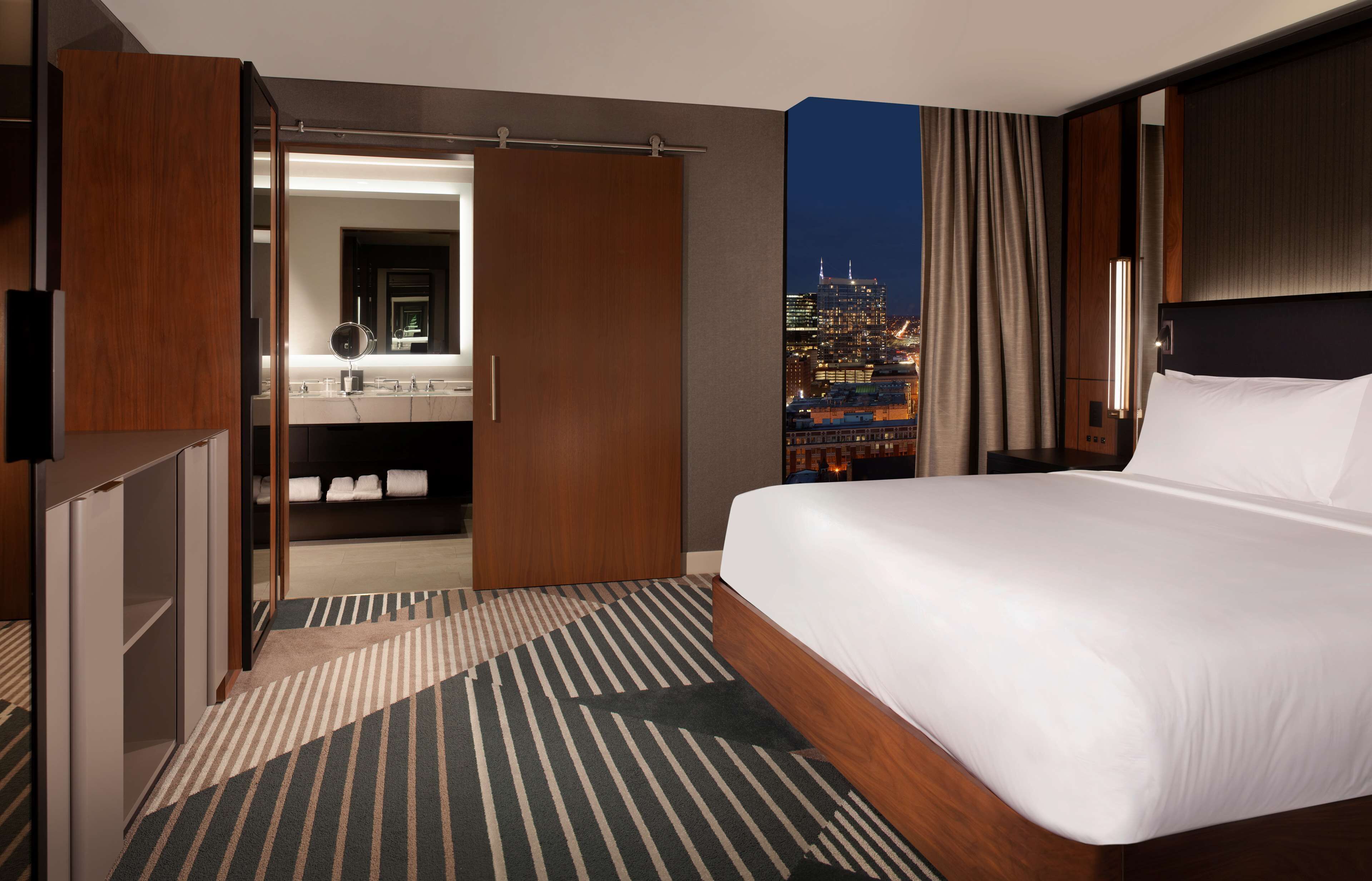 A hotel room with a large bed, a walk-in closet, a bathroom with a mirror, and a cityscape view at night.